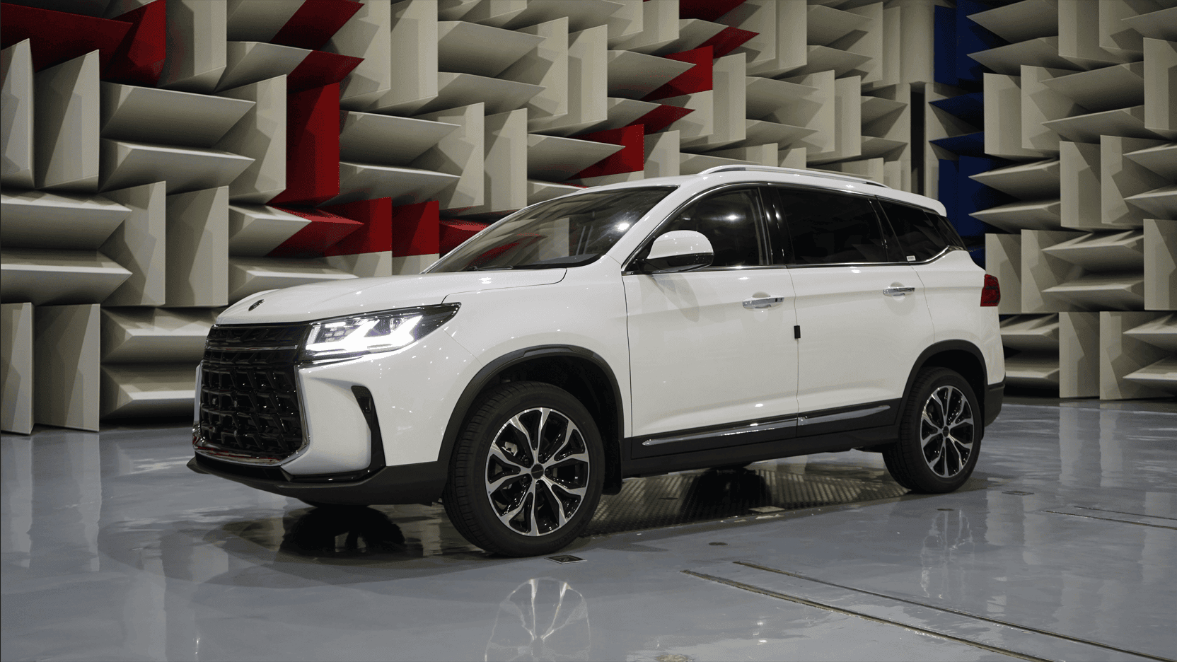 White SUV parked in an anechoic test chamber lined with acoustic foam panels and red/blue accents in the walls.
