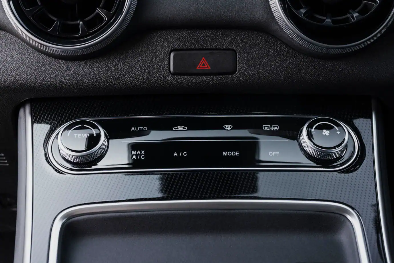 Close-up of a car's climate control panel with left temperature dial, central controls, hazard button above, and right fan dial on a glossy trim